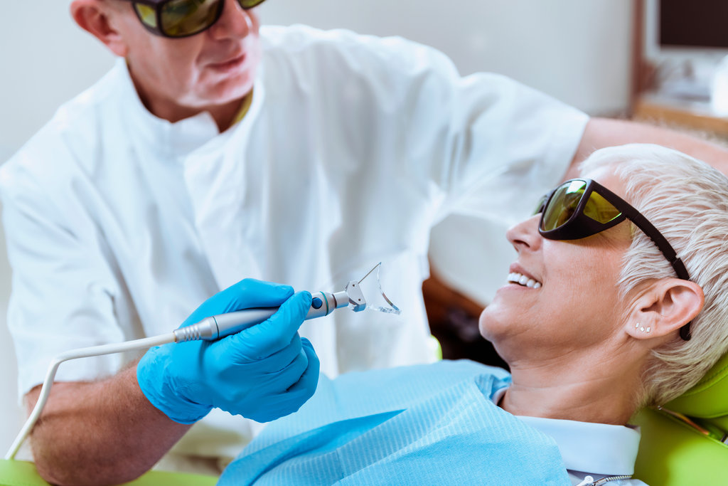 Laser Teeth Whitening. Elderly woman with short gray hair smiling and sitting in a dentist chair doing teeth whitening procedure. Doctor and patient wearing safety goggles.