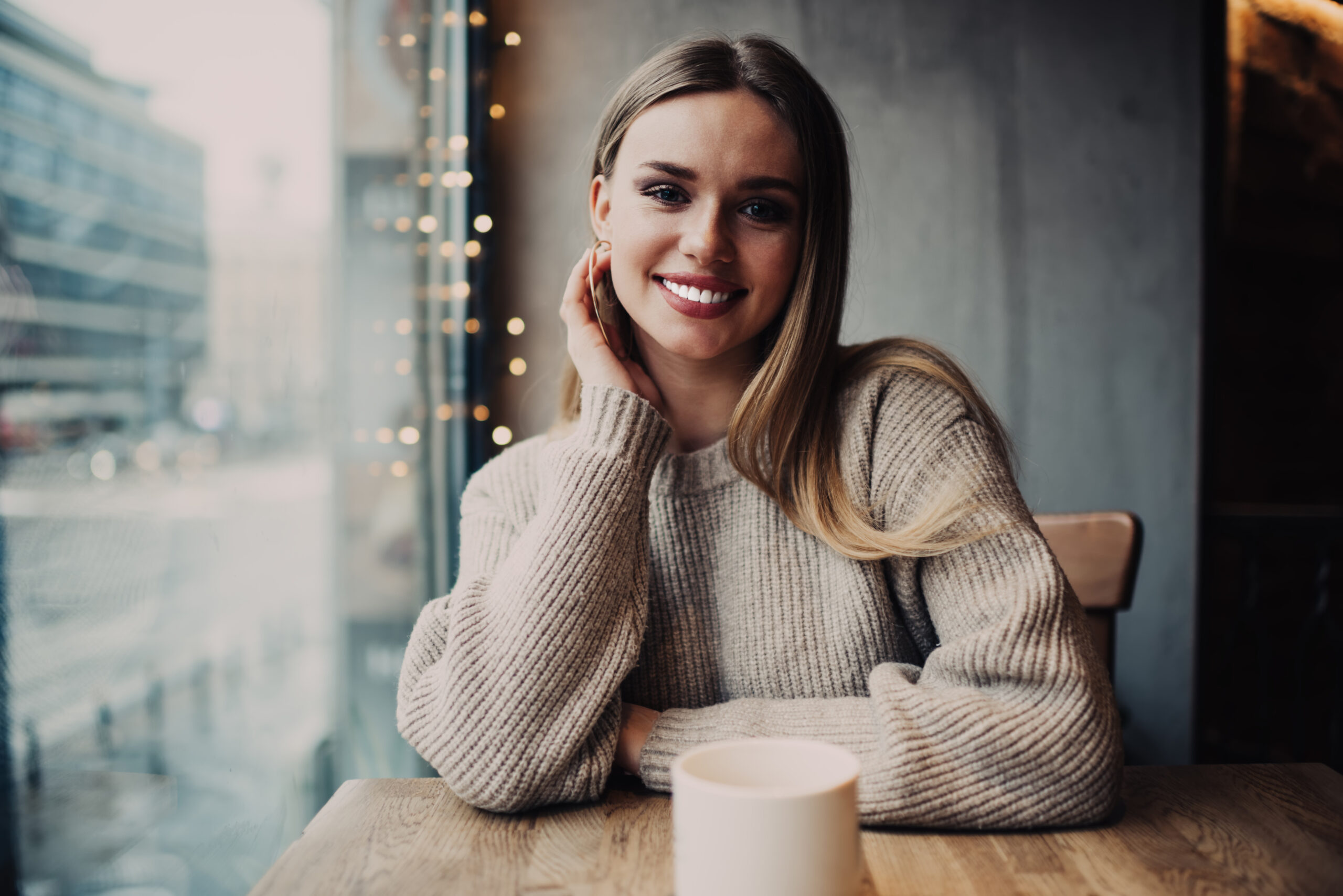 Half length portrait of cheerful female customer with perfect teeth smiling at camera during tea warming in cafe interior, happy Caucasian model recreating in cosy coffee shop enjoying leisure